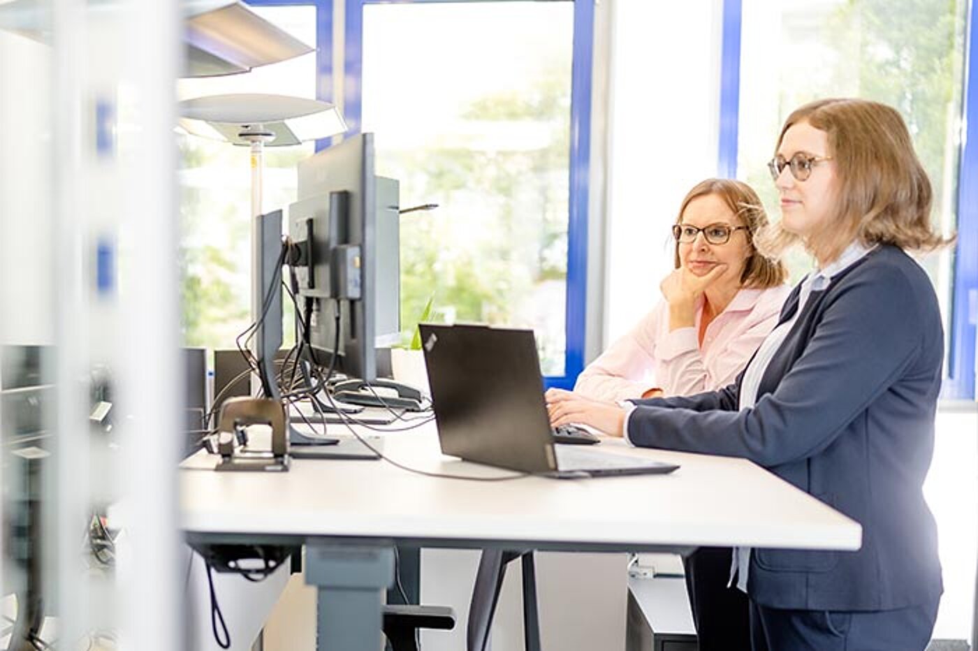 Zwei Frauen arbeiten an einem höhenverstellbaren Schreibtisch mit Monitoren und einem Laptop. Sie stehen nebeneinander und blicken auf die Bildschirme.