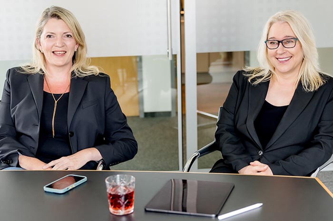 Zwei Frauen sitzen an einem Tisch und lächeln. Auf dem Tisch liegen ein Smartphone, ein Tablet und ein Glas mit Getränk.