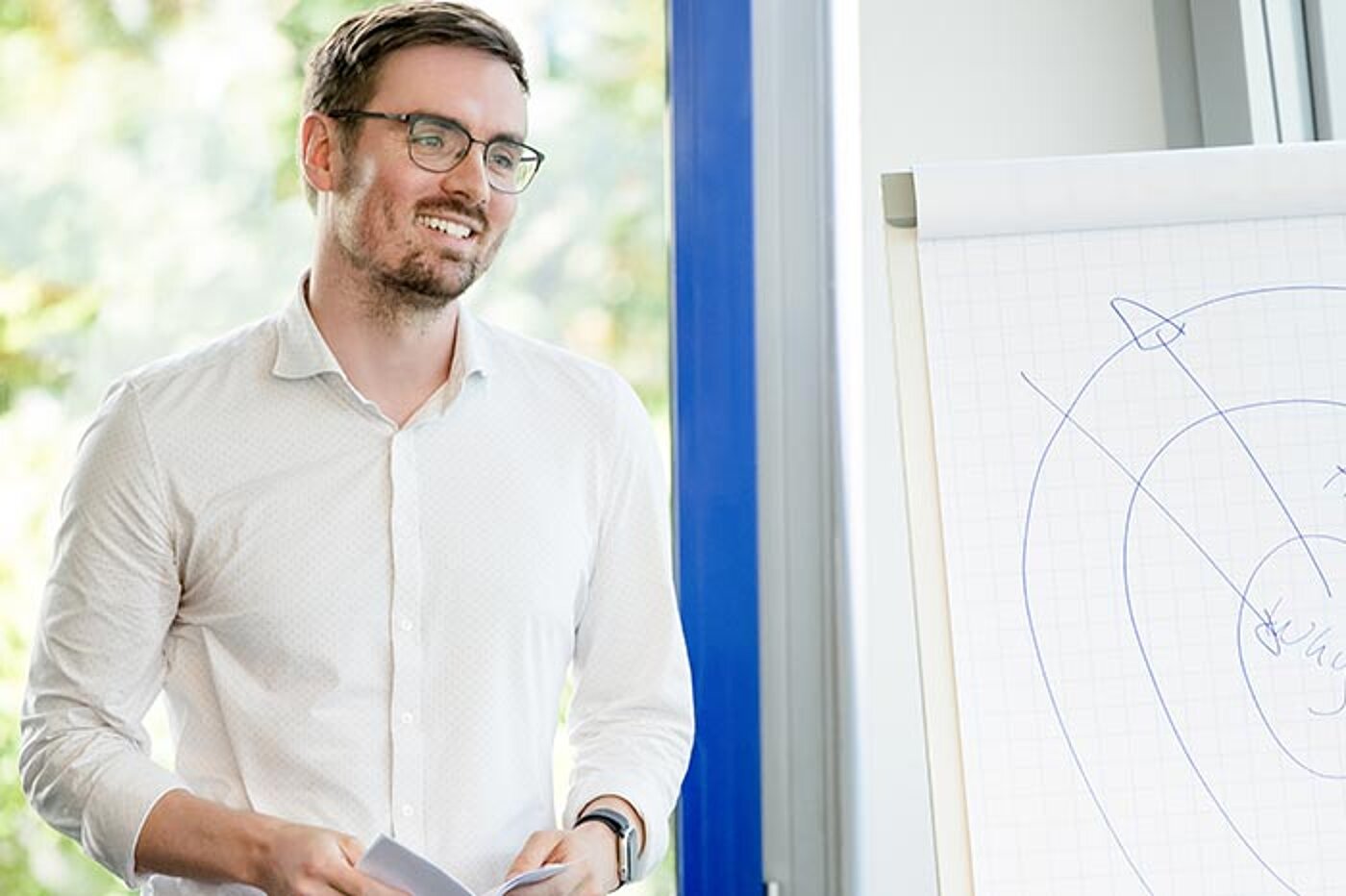 Ein Mann steht lächelnd neben einem Flipchart während eines Workshops. Auf dem Flipchart sind handgezeichnete Skizzen und Linien zu sehen.