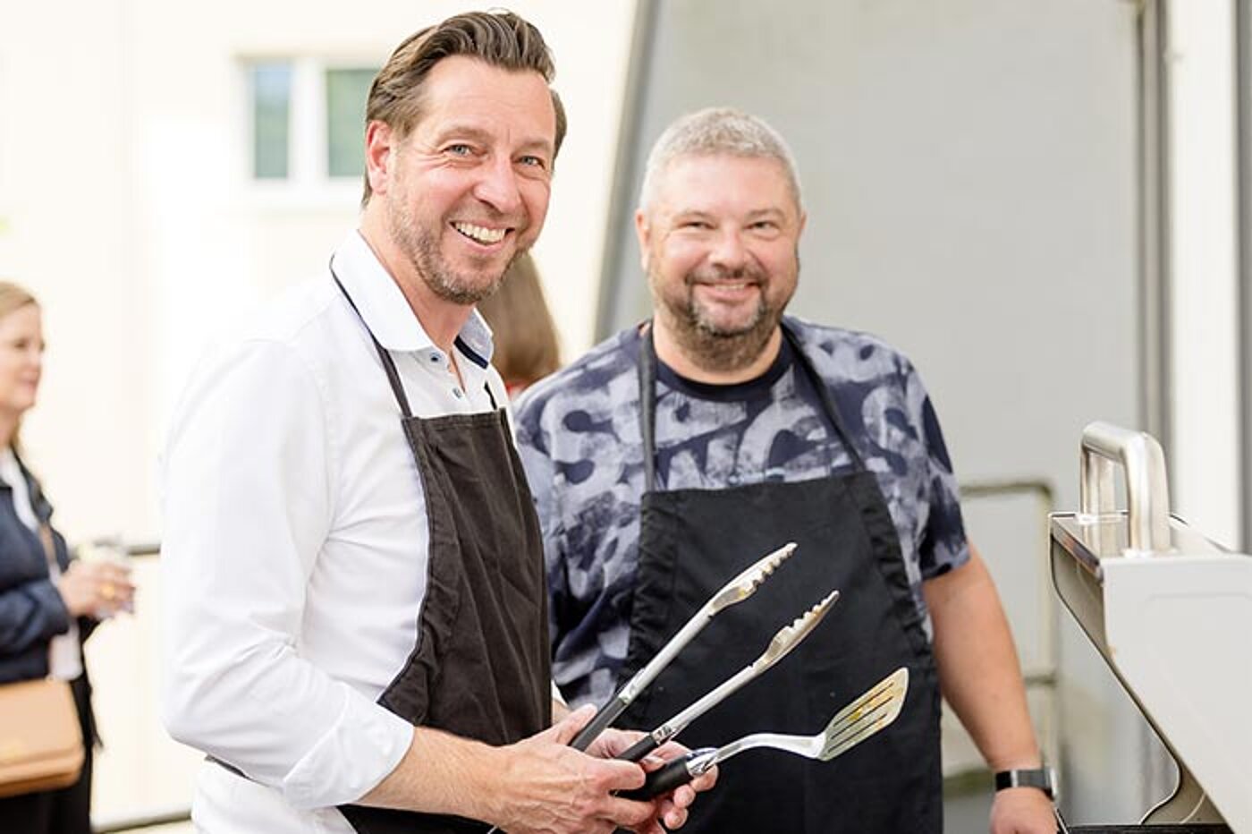 Zwei lächelnde Männer tragen Schürzen und stehen an einem Grill. Einer hält eine Grillzange und einen Pfannenwender in der Hand.