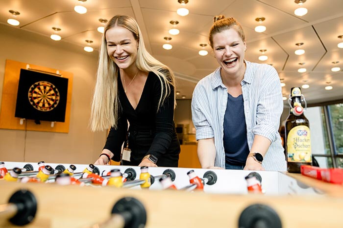 Zwei Frauen stehen an einem Tischkicker und lachen während des Spiels. Auf dem Tisch steht eine Radler-Bierflasche.