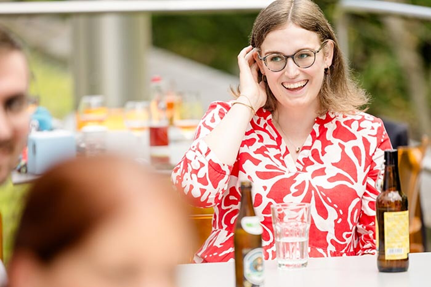 Eine lächelnde Frau mit Brille sitzt an einem Tisch im Freien. Auf dem Tisch stehen Bierflaschen und Gläser.