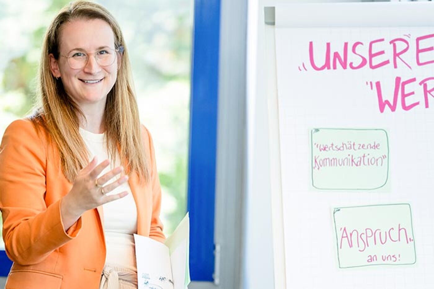 Eine lächelnde Frau steht neben einem Flipchart und gestikuliert während eines Workshops. Auf dem Flipchart sind handschriftliche Stichworte zu sehen.