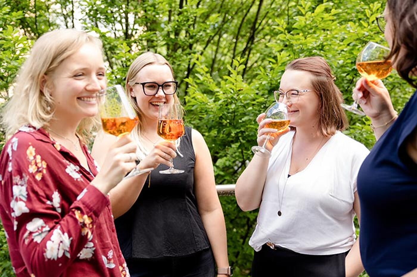 Vier Frauen stehen im Freien zusammen und halten Gläser mit Aperol Spritz in der Hand. Sie lächeln einander an.