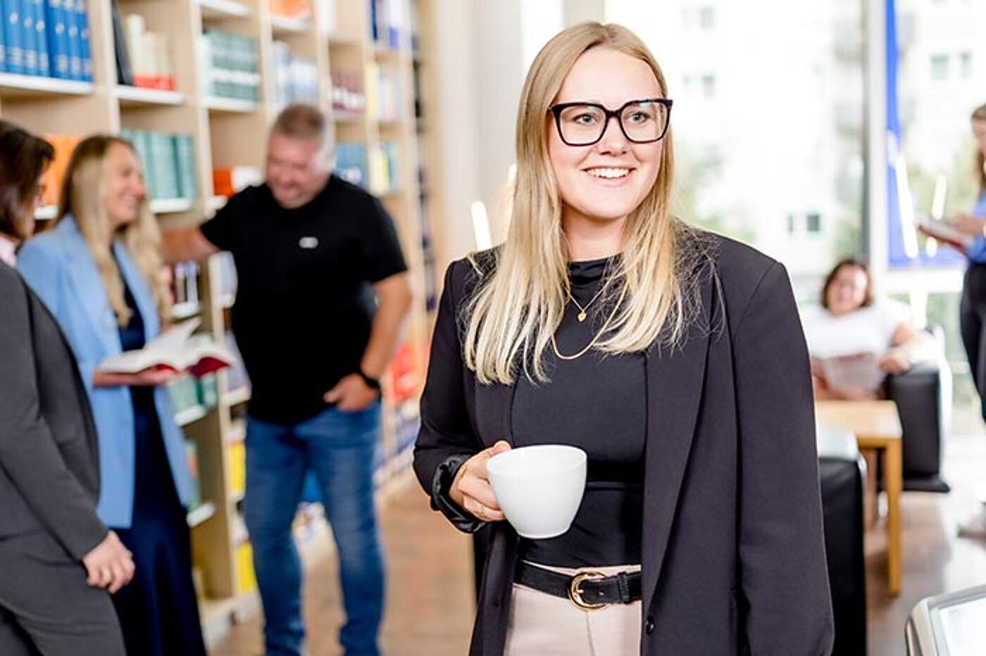 Eine lächelnde Frau steht in einem Raum mit Bücherregalen und hält eine Tasse in der Hand. Im Hintergrund sind weitere Personen zu sehen, die sich unterhalten oder lesen.